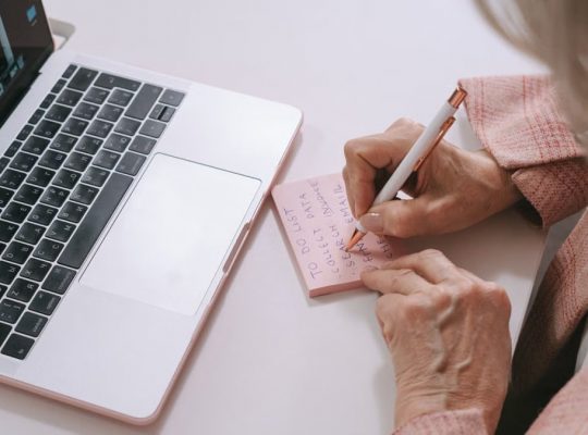 Close-up of a woman writing on a pink sticky note beside a laptop. Productive work setting.