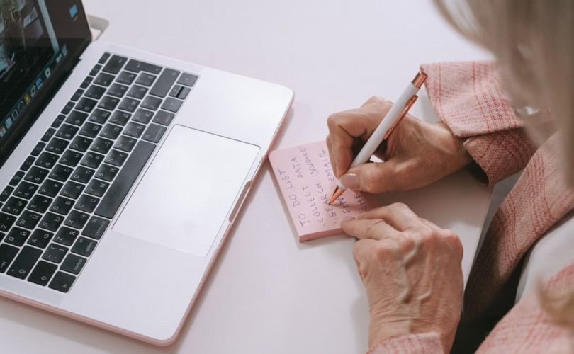 Close-up of a woman writing on a pink sticky note beside a laptop. Productive work setting.